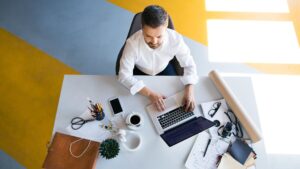 Homme travaillant sur un ordinateur portable dans un bureau lumineux, entouré de carnets, stylos et accessoires de travail.