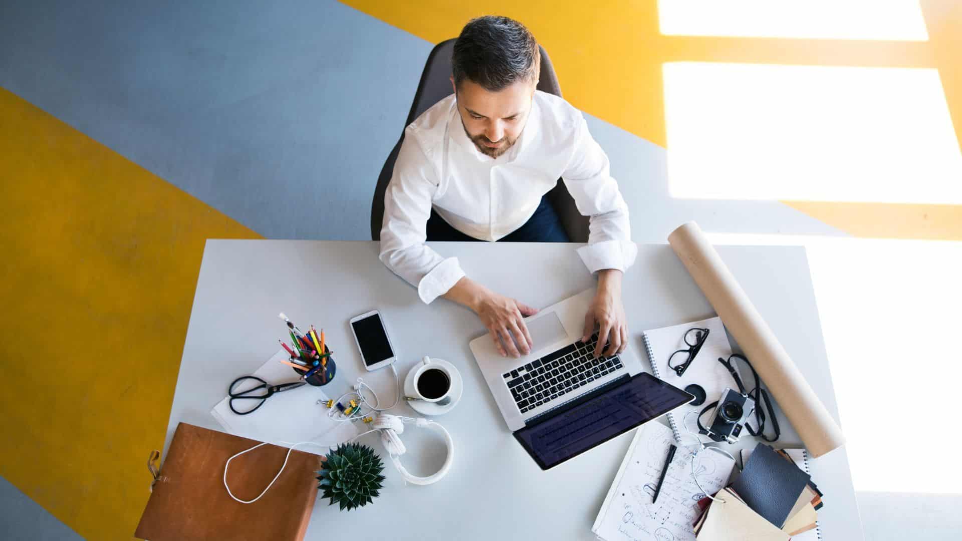 Homme travaillant sur un ordinateur portable dans un bureau lumineux, entouré de carnets, stylos et accessoires de travail.