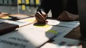 Une personne écrit avec un stylo sur des documents financiers étalés sur une table en bois, entourée de graphiques, post-its et d'une calculatrice, sous une lumière vive.